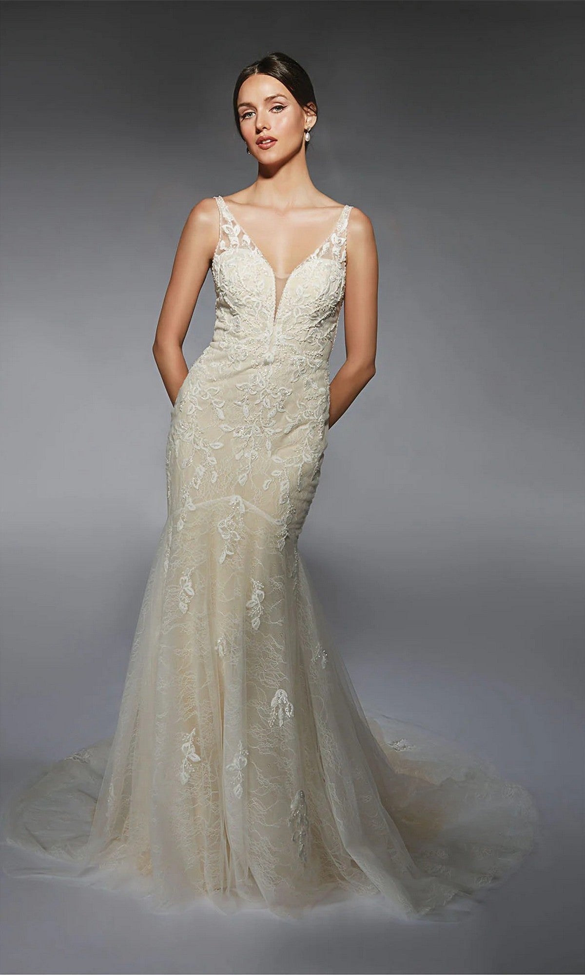 A woman poses in the Long White Dress: Alyce 7119, an ivory beaded lace mermaid gown with a deep V neckline and sheer straps, set against a plain gray background.